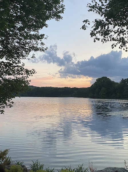 A lake under a sun-setting sky. There are trees around the water. The sky is blue and orange and yellow and has clouds in it. 