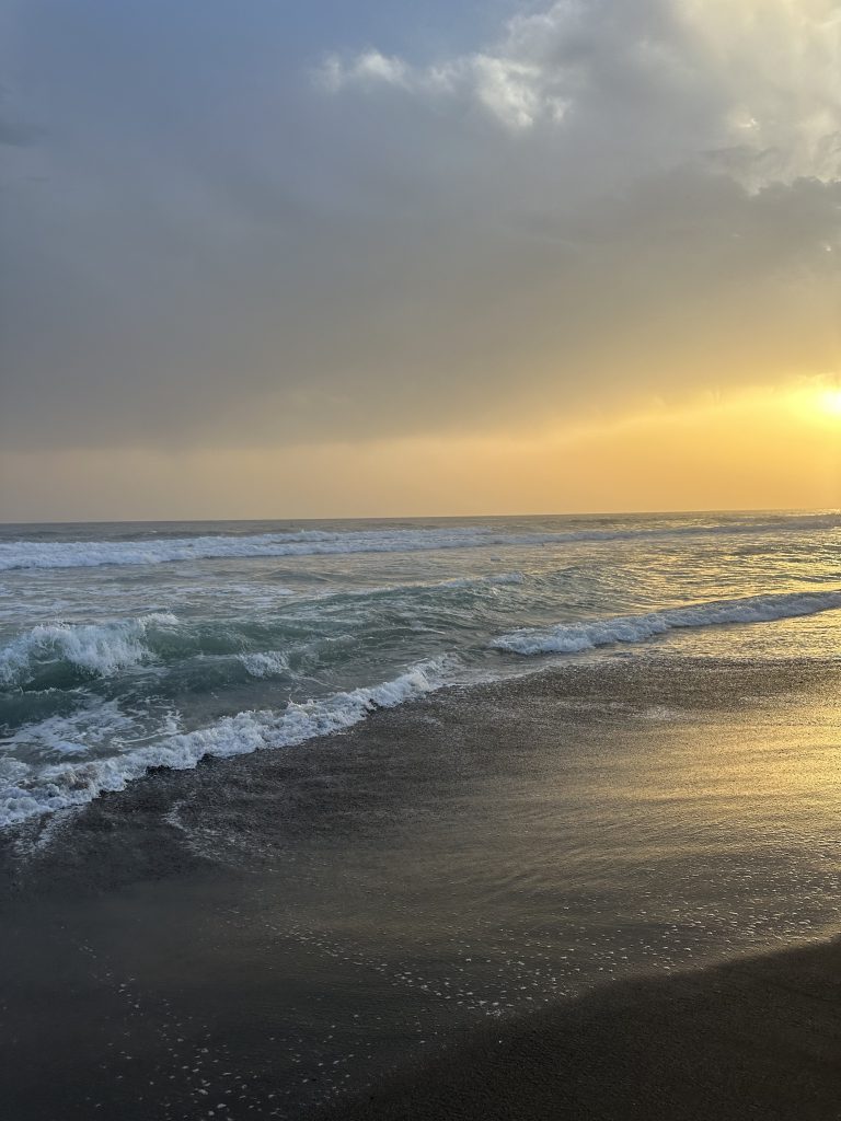 A beach with the sunset in the background. There are clouds over the sun as light gets through under the gray and makes the sky tinted yellow. 