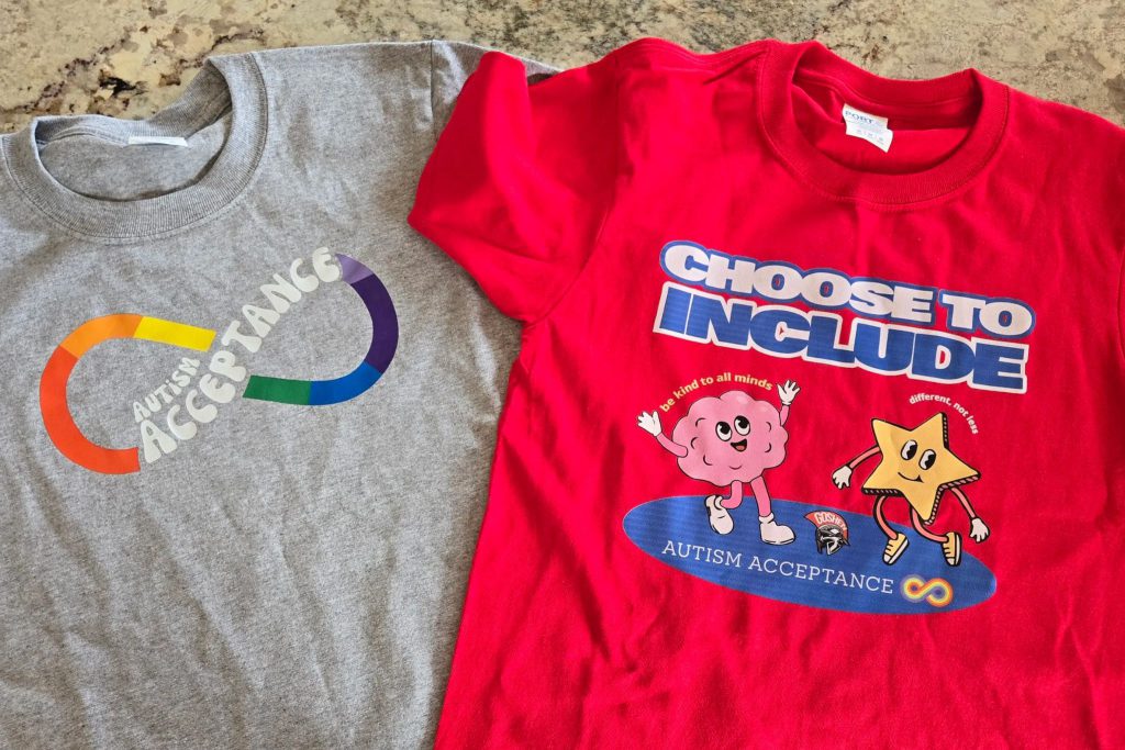 Two shirts are on a table and the picture is looking directly down at them. One on the left is gray and says Autism acceptance with a rainbow in an infinity swirl. The other says Choose to include and features a brain with a face and the words be kind to all minds above it. There is a star with arms and legs and a face and the words different, not less above it. The words autism acceptance are on a blue oval the two are standing on with the Goshen Gladiators logo and a rainbow infinity sign. 